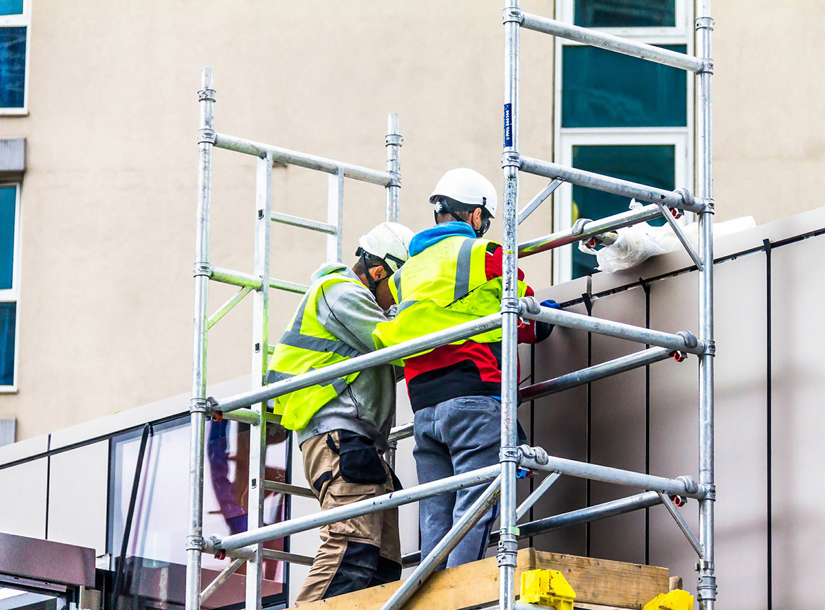 Two builders in retro-reflective clothing and helmets on scaffolding are engaged in repair work